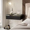 A modern bedroom scene featuring a floating black nightstand with gold trim, decorated with a bust, toiletries, and jewelry. A pair of black high heels are on the floor below. Produktbild-Vorschau