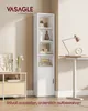 A tall, narrow white bookshelf with several open shelves and a closed cabinet at the bottom, placed in a room next to a desk with a lamp and a clock. A smaller wooden cabinet with drawers is visible on the left. Produktbild-Vorschau