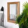 A rectangular mirror with a wooden frame hangs on a white wall, next to a black floor lamp and a potted plant. Produktbild-Vorschau