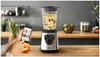 A person is using a smartphone to interact with a blender filled with fruit, suggesting a recipe or usage guide. Voir les images du produit