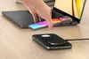 A hand is placing a smartphone onto a laptop, while a wireless charging pad sits on a wooden desk. Voir les images du produit