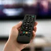 A hand holding a black TV remote control in front of a blurred television screen. Náhled obrázku produktu