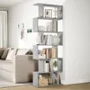 A gray, multi-tiered bookshelf filled with books and decorative items, placed in a living room next to a sofa and curtains. Náhľad obrázku produktu