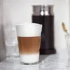 A layered coffee drink with foam in a glass, placed on a marble countertop. In the background is a frothing machine and some glasses. Náhľad obrázku produktu