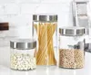 Three glass storage jars with silver lids are arranged on a white countertop in front of a white brick wall. The jars contain dried beans, spaghetti, and chickpeas. Produktbild-Vorschau