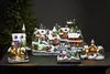 A collection of illuminated Christmas village houses and a church, set against a dark background with a blurred Christmas tree on the left. Podgląd zdjęcia produktu