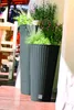Two tall, dark grey woven planters filled with green plants and some small purple flowers, placed on a wooden deck next to a white wall. A wooden chair and table are visible in the background. Produktbild-Vorschau