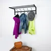 A black metal coat rack mounted on a white wall, with several items of clothing and a hat hanging on it. Below the coat rack, on a wooden crate, is a brown leather satchel. Produktbild-Vorschau