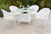 A white wicker patio set with a glass-topped table and four chairs is set up on a stone patio, surrounded by garden flowers. Produktbild-Vorschau