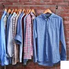 A rack of men's shirts hanging on wooden hangers against a brick wall. Produktbild-Vorschau