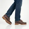 A person wearing jeans and brown leather boat shoes, shown from the knees down, against a white background. Anteprima dell’immagine prodotto