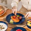 A person is cutting a piece of salmon on a blue plate with a floral pattern, accompanied by vegetables and lemons. Other dishes, including soup and a fruit bowl, are also visible. Náhled obrázku produktu