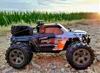 A remote-controlled monster truck is parked on a dirt path with a field of green plants in the background and the sun setting. Produktbild-Vorschau