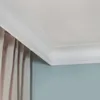 A close-up view of white crown molding installed at the junction of a ceiling and a light blue wall, with a beige curtain visible on the left. Produktbild-Vorschau