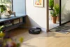 A black robot vacuum cleaner is docked in a corner of a room with wooden floors, near a doorway leading to a patio with plants. Produktbild-Vorschau
