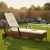 A rattan chaise lounge chair with a cream cushion is set up on a green lawn in a backyard garden, next to a small water feature and surrounded by flowers and a wooden fence. Náhled obrázku produktu