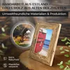 A wooden picture frame with a photo of a red boat on a beach is displayed on a wooden surface, with a circular inset image of a rustic cabin in the background. Text overlays highlight craftsmanship and eco-friendly materials. Produktbild-Vorschau