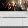 A white soundbar is placed on a white cabinet below a television displaying a concert. Produktbild-Vorschau