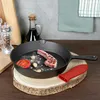 A cast iron frying pan with a lamb chop, tomato, and garlic inside, placed on a wooden board. A red silicone pot holder is next to the pan. In the background, there is a bottle of oil, a pepper grinder, and kitchen towels. Produktbild-Vorschau