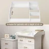 A product image showing a white changing table topper with shelves on the right, placed on top of a white dresser. The top part of the image shows a close-up of the topper with a measurement indicator, and the bottom part shows the topper in use with a bab Produktbild-Vorschau