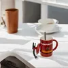 A red mug with a firefighter theme sits on a white table, with a brown cup and a white teacup in the background. Produktbild-Vorschau