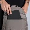 A person is putting a black notebook into the front pocket of a brown backpack. Anteprima dell’immagine prodotto