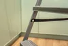 A close-up view of a gray metal step ladder, showing a black strap attached to one of the legs with a rivet. The background consists of light green paneled walls and a wooden floor. Anteprima dell’immagine prodotto