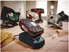 A collection of power tools, including a jigsaw, drill, and circular saw, are arranged on a wooden surface, with a battery charger and battery in the foreground. Produktbild-Vorschau