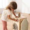 A young girl is playing with a wooden toy washing machine, ironing board, and iron in a bright, modern room. Produktbild-Vorschau