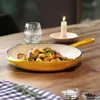 A yellow cast iron pan filled with roasted potatoes and herbs sits on a wooden table, next to a glass of water and a lit candle. Produktbild-Vorschau