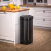 A black sensor trash can is placed next to a kitchen counter with a pitcher of orange juice and oranges. Náhled obrázku produktu