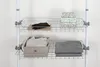 Two wire shelves are shown, each holding storage items. The top shelf has a dark item on a wooden board and three stacked boxes with patterns. The bottom shelf has a patterned box with folded fabric and a stack of folded gray fabric. Produktbild-Vorschau