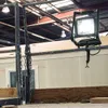 A work light is clamped to a metal bar in what appears to be a workshop or construction site. Podgląd zdjęcia produktu