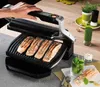 A hand is shown closing a Tefal Optigrill+ Elektrogrill GC718D10 with four pieces of salmon on the grill plates. The grill is on a wooden table with some side dishes and condiments. Voir les images du produit