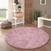 A round pink shaggy rug is placed on a wooden floor in a living room setting, next to a ladder shelf and a white console table. Produktbild-Vorschau