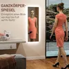 A woman in a coral dress walks past a full-length mirror with LED lighting. The mirror reflects another woman in the same dress. Text in German describes the mirror's features. Produktbild-Vorschau