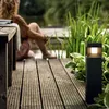 A person sits on a wooden deck next to a body of water, with a modern black outdoor lamp illuminating the scene. Anteprima dell’immagine prodotto