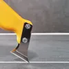 A close-up of a yellow and silver tool with a sharp blade, positioned on a gray tiled surface with a white grout line. Produktbild-Vorschau