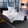 A bed with white bedding and pillows in a room with large windows overlooking a city at night. Produktbild-Vorschau