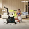A woman is demonstrating the features of a stationary bike in a living room setting. Produktbild-Vorschau
