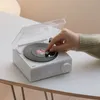 A hand is shown adjusting a knob on a white retro-style Bluetooth speaker designed to look like a record player. The speaker has a digital display showing the time. Podgląd zdjęcia produktu
