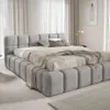A modern, upholstered bed in a neutral-toned bedroom with sheer curtains in the background. Produktbild-Vorschau