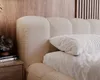 A close-up view of a beige boucle upholstered bed with a wooden headboard and a white pillow. Náhled obrázku produktu