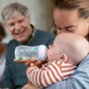 A woman is feeding a baby from a bottle, with a man smiling in the background. Produktbild-Vorschau