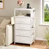 A white nightstand with four drawers and side pockets is placed next to a sofa in a living room setting. A phone is charging on top of the nightstand. Produktbild-Vorschau