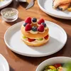 A close-up shot of a donut dessert topped with berries and cream, served on a white plate, with other food items and a small bowl of herbs in the background. Produktbild-Vorschau