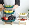 A person is holding a three-tiered fruit stand filled with various fruits, with another similar stand visible to the right. Náhled obrázku produktu