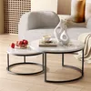 A set of two round nesting coffee tables with white marble-like tops and black metal bases are placed on a light wood floor. One table holds a bowl of red apples, and the other has a coffee cup and two decorative vases. A neutral-colored sofa is in the bac Voir les images du produit
