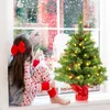 A young girl in pajamas sits on a windowsill, looking out at falling snow, with a small decorated Christmas tree next to her. Anteprima dell’immagine prodotto
