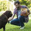 A man is crouching in a park, holding a bag of dog treats and interacting with his dog. Anteprima dell’immagine prodotto
